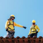 Two construction workers in safety gear discussing roofing project under clear blue sky.